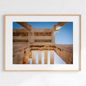 May include: Framed photograph of ancient Greek architecture. The image captures a low-angle view of weathered stone columns and beams against a bright blue sky. The composition highlights the architectural details and the play of light and shadow.