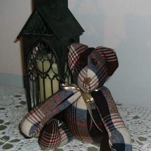 May include: A plaid teddy bear with a brown, blue, and white plaid pattern. The bear is sitting on a white crocheted tablecloth in front of a black metal lantern.