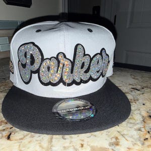 May include: White and black snapback hat with the name "Parker" in sparkly silver letters. The hat has a flat brim and a snap closure on the back. The hat is a stylish accessory.