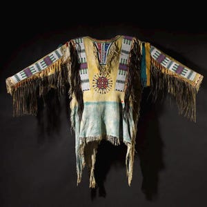 Puede incluir: Una camisa de estilo nativo americano con una combinación de colores amarillo, azul y crema. La camisa presenta intrincados abalorios en varios colores y largas flecos a lo largo de las mangas y el dobladillo. Un diseño circular está centrado en el pecho.