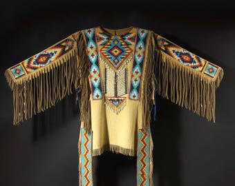 May include: A tan Native American shirt with detailed beadwork in red, blue, yellow, and white. The shirt has long fringe on the sleeves and hem, and geometric patterns. The design includes a central diamond motif and vertical bead stripes.