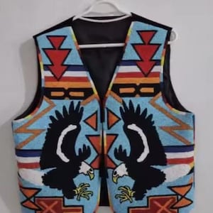 Puede incluir: Un chaleco con cuentas con un reverso negro, colgado de una percha blanca. El dise&ntilde;o presenta dos &aacute;guilas negras sobre un fondo azul claro. Los patrones geom&eacute;tricos en rojo, naranja y blanco completan el dise&ntilde;o.