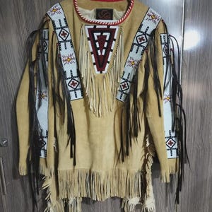 May include: Tan suede shirt with intricate beadwork and fringe detailing. The shirt features a geometric design in red, white, and black on the front and sleeves. Black fringe hangs from the sleeves and hem, adding movement and texture to the garment.