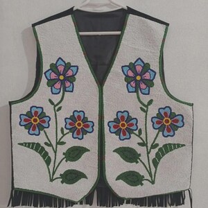 Puede incluir: Un chaleco blanco con cuentas con la espalda y el ribete negros, adornado con diseños florales coloridos. Las flores son azules, rosas y rojas, con hojas y tallos verdes. El chaleco tiene flecos en la parte inferior y está colgado de una percha blanca.