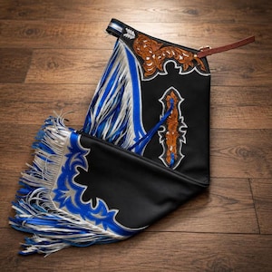 May include: Black leather chaps featuring intricate brown floral tooling at the top and centre. The garment is accented with vibrant blue and silver leather overlays, long silver and blue fringe along the side, and a brown leather adjustable strap.