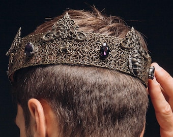 Puede incluir: Corona dorada antigua adornada con piedras preciosas moradas. La corona se lleva en la cabeza. También se ven un collar y pulseras plateadas. El fondo es negro.