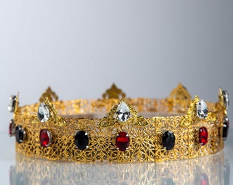 Op de afbeelding: Een goudkleurige kroon met ingewikkelde filigraan details. De kroon is versierd met rode, zwarte en heldere edelstenen. Het ontwerp heeft een reeks decoratieve punten langs de bovenrand, met een weerspiegeling.