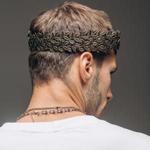 Puede incluir: Una diadema de corona de laurel en tono bronce met&aacute;lico con un patr&oacute;n detallado de hojas en doble fila. El accesorio se coloca alrededor de la cabeza, descansando sobre las orejas, y presenta un acabado texturizado que imita el follaje.