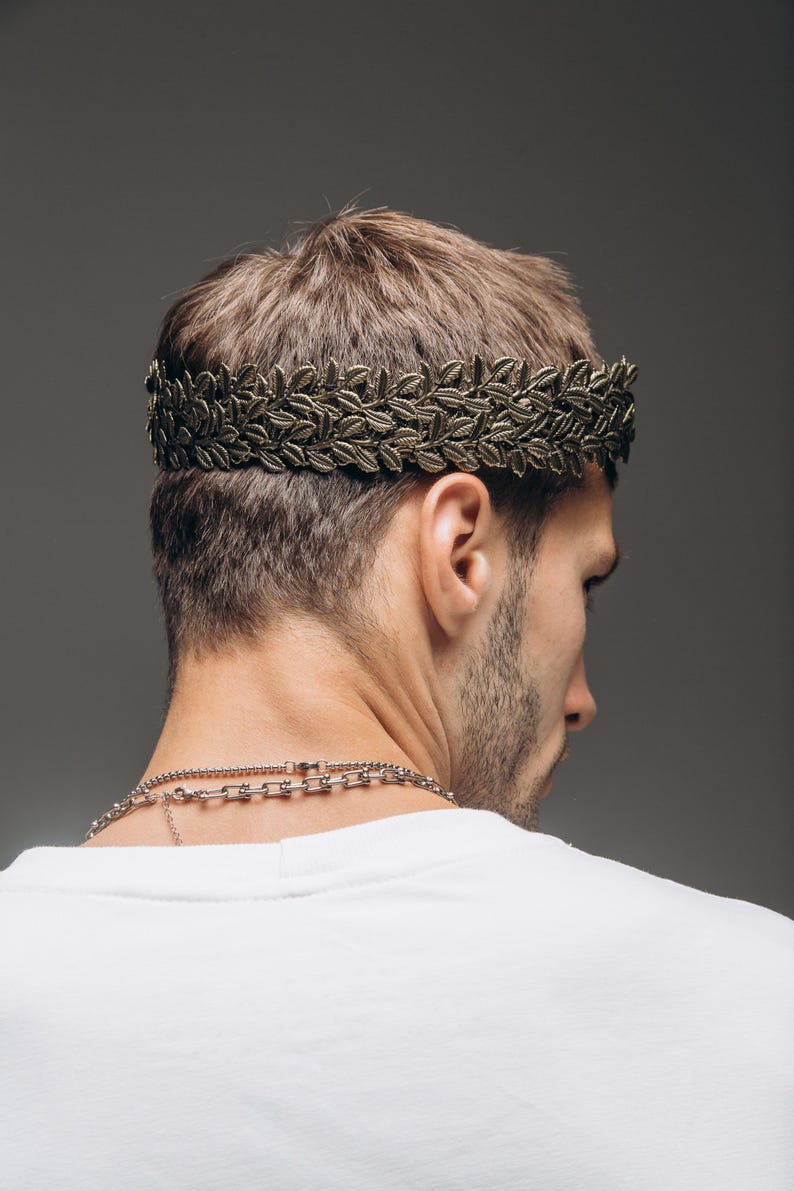 Puede incluir: Una diadema de corona de laurel en tono bronce met&aacute;lico con un patr&oacute;n detallado de hojas en doble fila. El accesorio se coloca alrededor de la cabeza, descansando sobre las orejas, y presenta un acabado texturizado que imita el follaje.