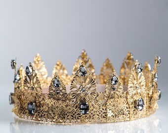 Op de afbeelding: Een goudkleurige kroon met ingewikkelde filigraan details en versierd met donkergrijze edelstenen. De kroon heeft een reeks puntige bogen en een cirkelvormige basis. Een koninklijk accessoire voor speciale gelegenheden.