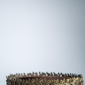 Puede incluir: Una corona de hojas doradas, probablemente de metal, se muestra sobre un fondo blanco. La corona presenta un dise&ntilde;o detallado de hojas alrededor de toda la circunferencia. Las hojas est&aacute;n muy juntas, creando una apariencia texturizada. Adecuada para eventos tem&aacute;ticos.