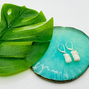 Puede incluir: Un par de pendientes blancos de piedra apilada con cierres de palanca plateados. Los pendientes se exhiben sobre un posavasos decorativo redondo de color turquesa, junto a una gran hoja verde.