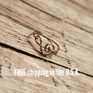 May include: A gold-colored ring in the shape of a treble clef. The ring is made of thin wire and sits on a wooden surface. The ring is a musical-themed jewelry item.