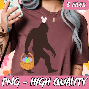 Puede incluir: Una camiseta de color marrón rojizo con un gráfico de un Bigfoot con orejas de conejo y llevando una cesta de huevos de Pascua coloridos. El texto "LOVE" es visible en la manga de la camiseta.