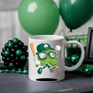 May include: White ceramic mug featuring a green shamrock cartoon character wearing sunglasses and a baseball cap, holding a baseball bat and a baseball. The shamrock is sporting a green and white baseball uniform. The mug has a black handle.