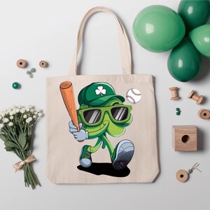 May include: A white canvas tote bag featuring a green shamrock character wearing a baseball cap, sunglasses, and holding a baseball bat. The shamrock is holding a baseball in its hand. The bag has a green shamrock design on the front.