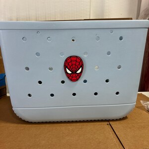 May include: A light blue tote bag with multiple circular holes. A red and black Spider-Man face emblem is affixed to the front. The bag is made of a durable material and has a rounded bottom.
