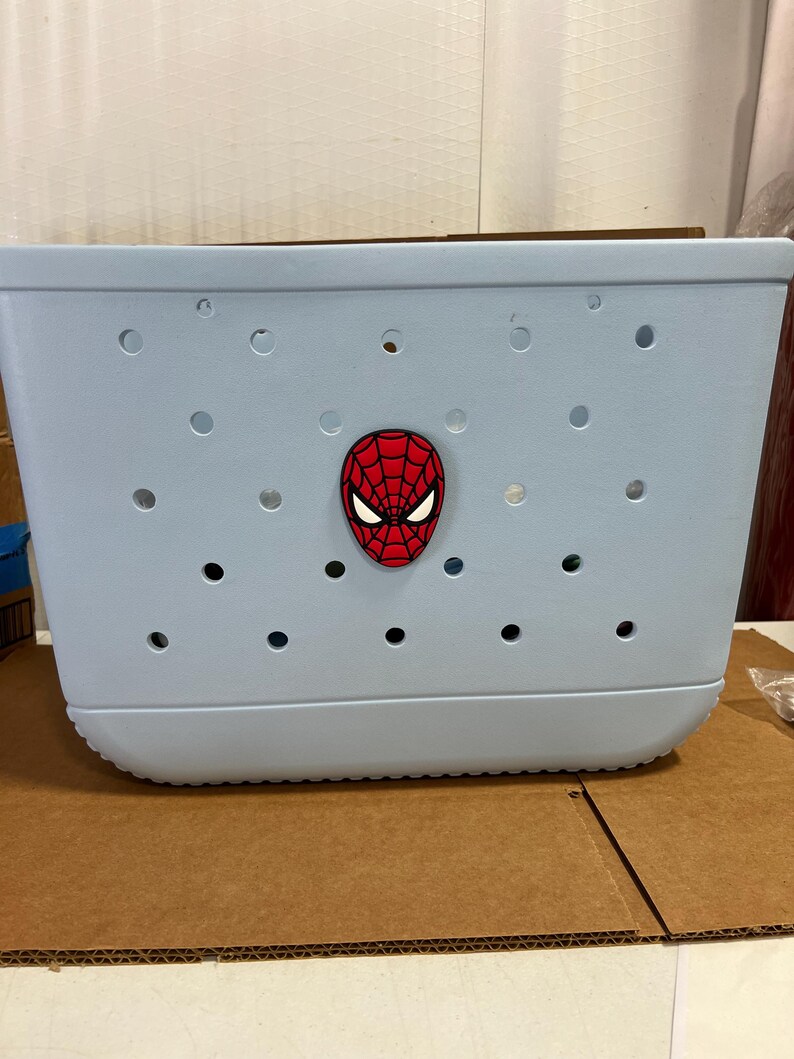 May include: A light blue tote bag with multiple circular holes. A red and black Spider-Man face emblem is affixed to the front. The bag is made of a durable material and has a rounded bottom.
