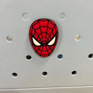 May include: A red and black Spider-Man face charm with white eye details. The charm is affixed to a light gray surface with several circular holes.