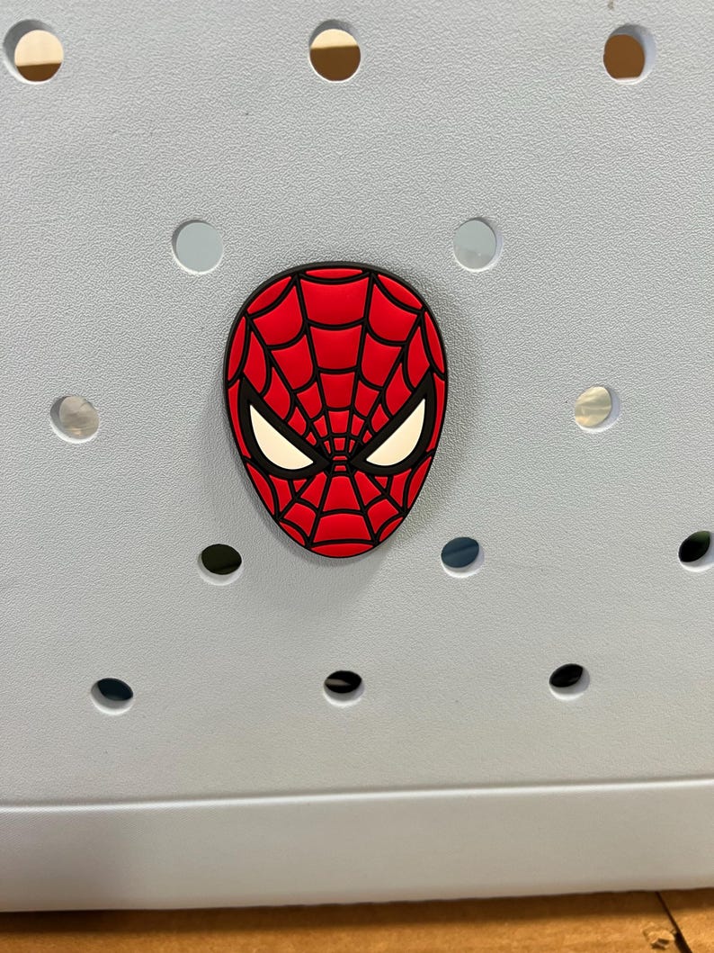 May include: A red and black Spider-Man face charm with white eye details. The charm is affixed to a light gray surface with several circular holes.