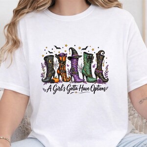 May include: A white short-sleeve t-shirt featuring a graphic print of five distinct boots in black, orange, purple, and green colors, accented with small pumpkins, bats, and stars. The text "A Girl's Gotta Have Options" is printed in black below the boots.