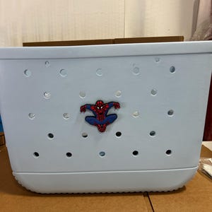 Peut inclure: Un sac fourre-tout rectangulaire bleu clair fabriqu&eacute; dans un mat&eacute;riau souple et perfor&eacute;. Le sac pr&eacute;sente une s&eacute;rie de trous circulaires r&eacute;guli&egrave;rement espac&eacute;s sur sa surface et un embl&egrave;me central en caoutchouc repr&eacute;sentant Spider-Man en rouge et bleu.