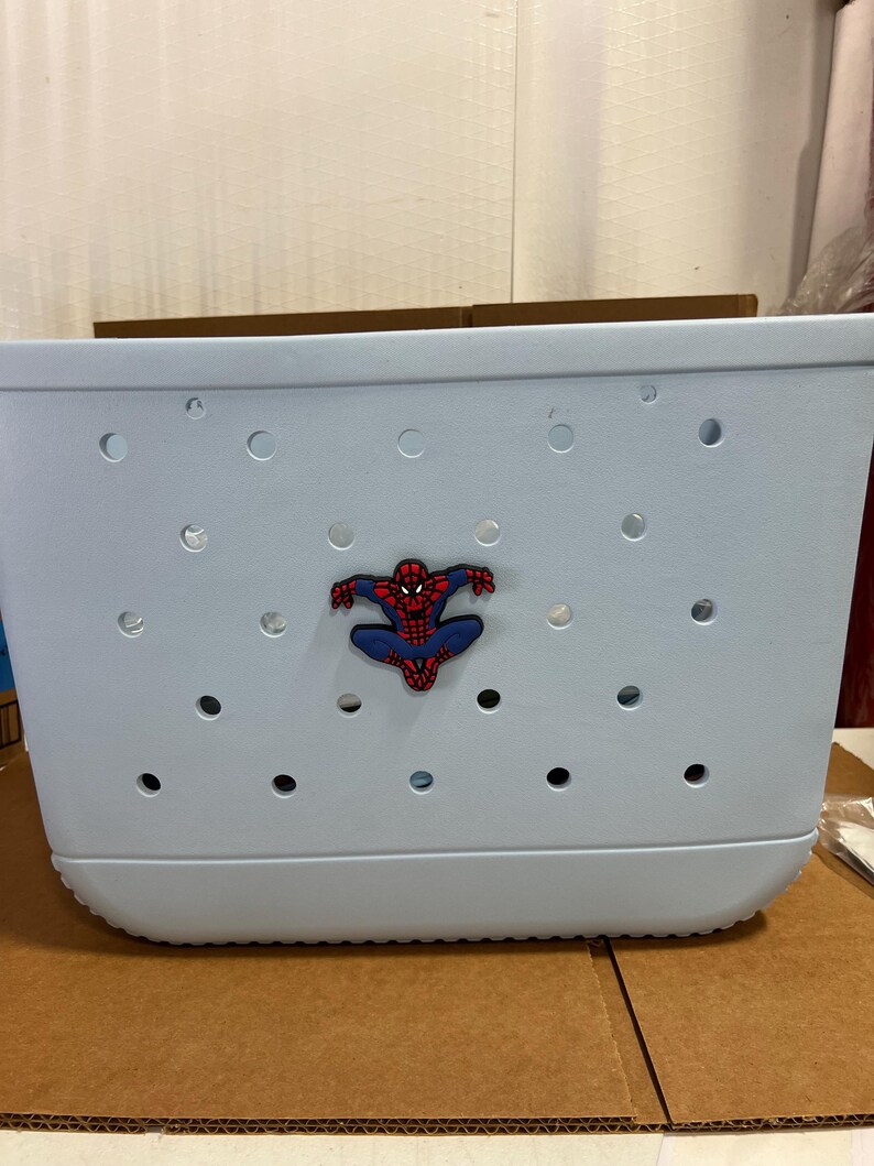 Peut inclure: Un sac fourre-tout rectangulaire bleu clair fabriqu&eacute; dans un mat&eacute;riau souple et perfor&eacute;. Le sac pr&eacute;sente une s&eacute;rie de trous circulaires r&eacute;guli&egrave;rement espac&eacute;s sur sa surface et un embl&egrave;me central en caoutchouc repr&eacute;sentant Spider-Man en rouge et bleu.