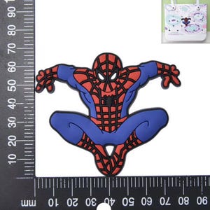 Peut inclure: Un embl&egrave;me en caoutchouc repr&eacute;sentant Spider-Man avec un costume rouge et bleu et des d&eacute;tails de toile noirs. La figurine est en position accroupie. Une r&egrave;gle noire est visible sur les bords gauche et inf&eacute;rieur, indiquant environ 60 mm.