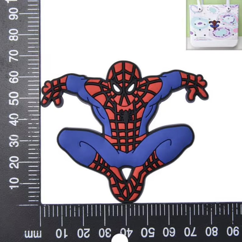 Peut inclure: Un embl&egrave;me en caoutchouc repr&eacute;sentant Spider-Man avec un costume rouge et bleu et des d&eacute;tails de toile noirs. La figurine est en position accroupie. Une r&egrave;gle noire est visible sur les bords gauche et inf&eacute;rieur, indiquant environ 60 mm.