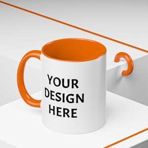 May include: White ceramic mug with an orange rim and handle. The mug has the text "YOUR DESIGN HERE" printed on the front.