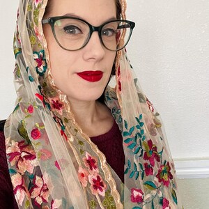 May include: A sheer beige scarf with floral embroidery in shades of pink, red, and green. The scarf has a scalloped edge and is draped over a person's shoulders.