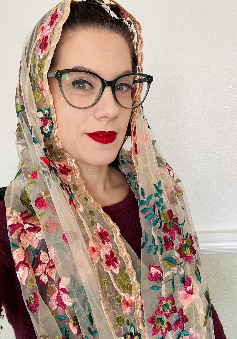 May include: A sheer beige scarf with floral embroidery in shades of pink, red, and green. The scarf has a scalloped edge and is draped over a person's shoulders.