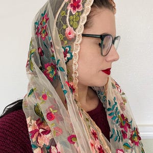 May include: A sheer beige shawl with floral embroidery in shades of pink, red, green, and teal. The shawl has a scalloped edge with a gold trim.