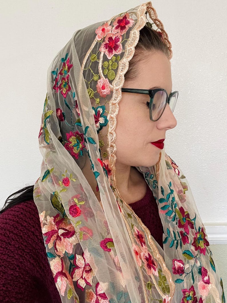 May include: A sheer beige shawl with floral embroidery in shades of pink, red, green, and teal. The shawl has a scalloped edge with a gold trim.