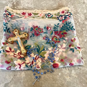 May include: A white sheer fabric with floral embroidery, a wooden cross with a silver figure of Jesus, and a blue rosary with a silver pendant.