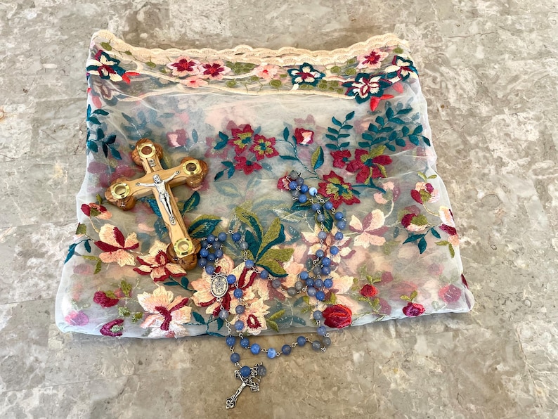 May include: A white sheer fabric with floral embroidery, a wooden cross with a silver figure of Jesus, and a blue rosary with a silver pendant.