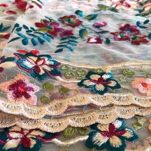 May include: White sheer fabric with a delicate floral embroidery pattern in shades of pink, green, and teal. The fabric has a scalloped edge.