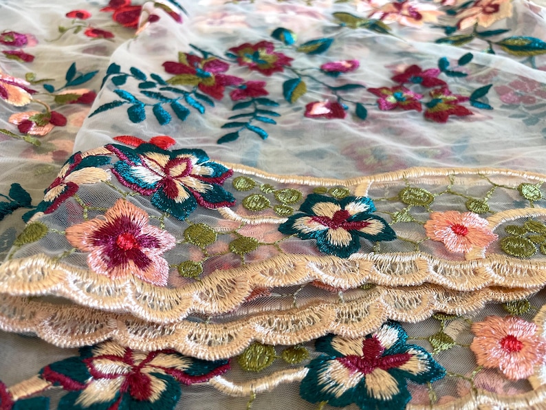 May include: White sheer fabric with a delicate floral embroidery pattern in shades of pink, green, and teal. The fabric has a scalloped edge.