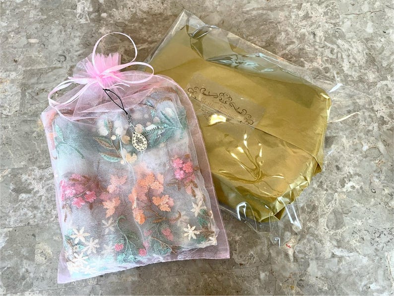 May include: A pink tulle bag with a floral embroidery design. The bag is tied with a ribbon and has a silver charm hanging from it.