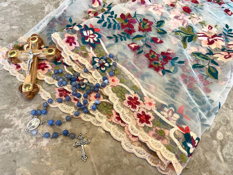 May include: A blue rosary with a silver crucifix and a silver medal of the Virgin Mary, lying on a white lace fabric with floral embroidery.