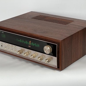 Pode incluir: Um receptor estéreo vintage com um gabinete de grão de madeira escura e uma placa frontal prateada. A placa frontal apresenta botões e um visor com números verdes iluminados. A parte superior do receptor tem ranhuras de ventilação horizontais.
