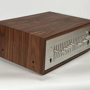 Può includere: Un equalizzatore audio vintage con un cabinet in legno di noce e un pannello frontale in metallo spazzolato. Il pannello frontale presenta numerosi cursori e manopole di controllo. L'unità è progettata per regolare le frequenze audio.