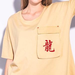 May include: Mustard yellow t-shirt with a pocket featuring red Japanese characters. The pocket is sewn onto the shirt with red stitching. The shirt has short sleeves and a crew neck.