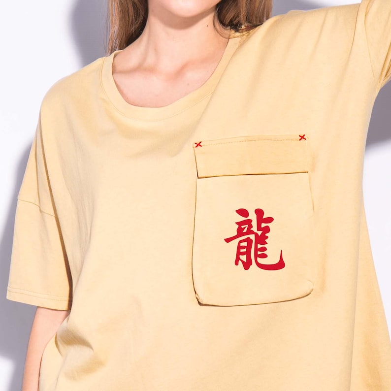 May include: Mustard yellow t-shirt with a pocket featuring red Japanese characters. The pocket is sewn onto the shirt with red stitching. The shirt has short sleeves and a crew neck.