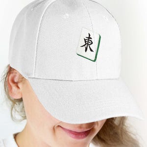 Puede incluir: Gorra de b&eacute;isbol blanca con un dise&ntilde;o de ficha de Mahjong. La ficha tiene un car&aacute;cter negro y un borde verde. La gorra est&aacute; hecha de una tela texturizada y tiene una visera curva. La gorra est&aacute; siendo usada.
