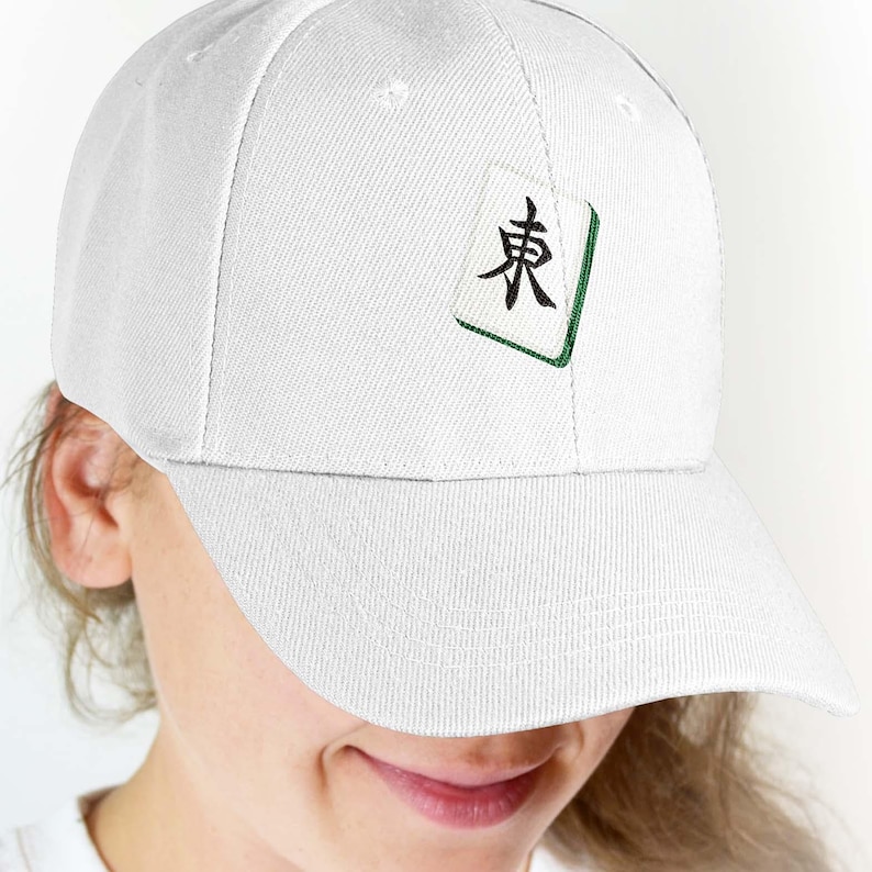 Puede incluir: Gorra de b&eacute;isbol blanca con un dise&ntilde;o de ficha de Mahjong. La ficha tiene un car&aacute;cter negro y un borde verde. La gorra est&aacute; hecha de una tela texturizada y tiene una visera curva. La gorra est&aacute; siendo usada.