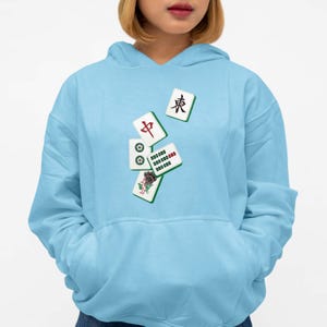 Puede incluir: Sudadera con capucha azul claro con un dise&ntilde;o de fichas de Mahjong. Las fichas son blancas con s&iacute;mbolos rojos, verdes y negros. La sudadera tiene un bolsillo delantero y una capucha con cord&oacute;n.