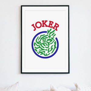 May include: Framed art print featuring the word "JOKER" in red above a green and blue graphic. The graphic is a stylized design within a blue circle, set against a white background. The artwork is framed in a black frame.