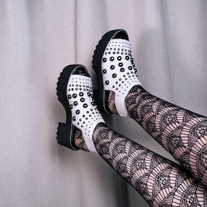 May include: A pair of white open-toe sandals featuring a thick black lug sole and a block heel. The upper part of the sandals is adorned with numerous small, round, black metal studs arranged in a pattern across the white material.