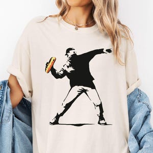 May include: Ivory-colored t-shirt featuring a black and white graphic of a person throwing a hot dog. The text "SunsetAndStitchCo" and "Ivory" are visible. The shirt is worn with a light blue denim jacket.